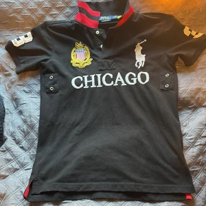 Svart pikétröja Polo Ralph Lauren - Svart pikétröja från Polo Ralph Lauren med broderad Chicago-logga, stort vitt polospelare-märke och siffran 3 på ryggen. Röd och svart krage, detaljer i gult och vitt samt broderad emblem på bröstet. Slim fit-modell i bomull, perfekt för dig som gillar sportig stil.