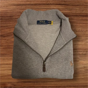Polo Ralph Lauren Quarter Zip Grå Storlek Large  - Snygg och klassisk grå Polo Ralph Lauren quarter zip i storlek L. Äkta produkt med broderad logga och läderdetalj på dragkedjan. Mjuk bomull, bekväm passform och i mycket bra skick utan defekter. 