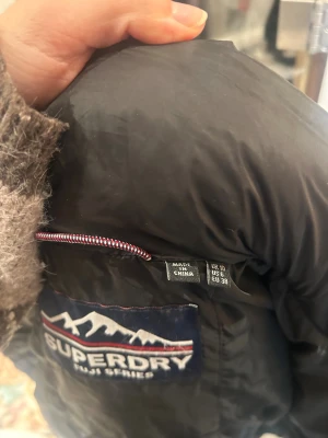 Svart pufferjacka Superdry  - Svart pufferjacka från Superdry Fuji Series med huva och dragkedja framtill. , två fickor med dragkedja och Superdry-logga på sidan, bara använd en gång