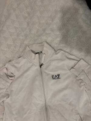 Vit EA7 zipjacka med hög krage - Säljer en fräsch vit zipjacka från EA7 Emporio Armani med stilren logga på bröstet. Tröjan har hög krage, hel dragkedja och två sidofickor. Materialet är mjukt och skönt , perfekt för chill och streetwear. Passformen är normal och ärmarna är långa.