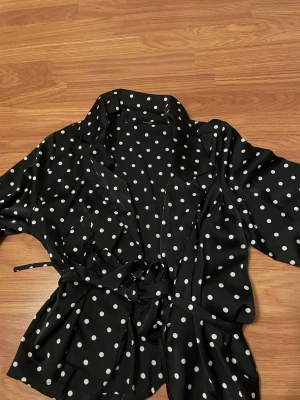 Svart prickig omlottblus med bälte - Svart omlottblus med vita prickar och knytband i midjan. Blus har klassisk krage, lång ärm och är tillverkad i ett mjukt, lätt material som ger ett snyggt fall. Perfekt för dig som gillar retrovibbar och vill sticka ut med ett lekfullt mönster.