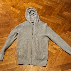 Grå stickad hoodie med dragkedja från All Saints - Säljer en grå stickad hoodie med huva och snörning. Tröjan har dragkedja framtill och två fickor. Perfekt för dig som gillar en chill och enkel stil. Långärmad och ribbad struktur ger extra skön känsla.