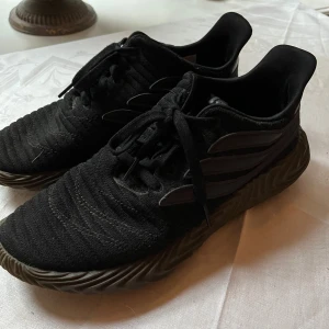 Adidas sneakers svarta - Svarta Adidas sneakers med stickad ovandel och klassiska tre ränder på sidan. Skorna har en unik vågig sula i mörkgrått och snörning framtill. Perfekta för dig som gillar sportig stil och vill ha något som sticker ut. Materialet är mjukt och följsamt för extra komfort.