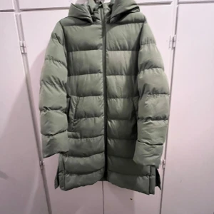 Grön lång pufferjacka med huva  - Snygg ljusgrön pufferjacka med huva och dragkedja framtill. Jackan är lång, har knappar i sidorna och breda, quiltade kanaler. Perfekt för kalla dagar och har en clean, modern look. Jackan är S men den passar M