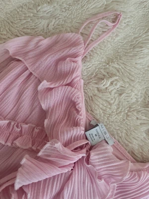 Ljusrosa volangtop från MISSGUIDED - Superfin ljusrosa linnetopp från MISSGUIDED med tunna axelband och volangdetaljer. Toppen har ribbad struktur, omlottknytning framtill och är gjord i ett lätt, transparent syntetmaterial. Perfekt för dig som gillar romantisk och söt stil.