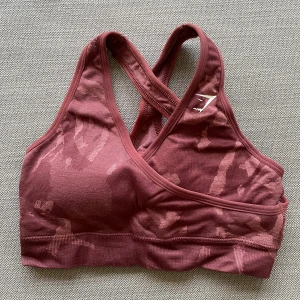 Vinröd sporttopp från Gymshark - Säljer en vinröd sporttopp från Gymshark med camouflagemönster och korsade axelband i ryggen. Knappt använd och därav i superbra skick.💕
