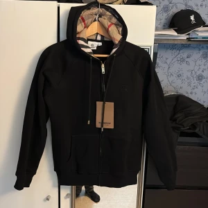 Sprillans ny Burberry. Säljs för att den var för liten. - Svart hoodie från Burberry med klassiskt rutigt foder i huvan, guldfärgade detaljer på snörningen och dragkedja framtill. Tröjan har känguruficka och diskret Burberry-logga på bröstet. Perfekt för dig som vill ha en stilren och lyxig look.