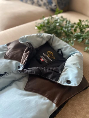 Canada Goose ljusblå dunväst med bruna detaljer - Snygg dunväst från Canada Goose i ljusblått med bruna axelpartier i skinn. Västen har hög krage, dragkedja och tryckknappar framtill samt två fickor med dragkedja. Perfekt för dig som vill ha en cool och varm look under höst och vinter.