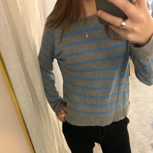 Grå och blå randig tröja - Snygg grå tröja med blå horisontella ränder. Modellen har rund hals och långa ärmar, perfekt för dig som gillar enkel men stilren design. Materialet känns mjukt och lätt, vilket gör den skön att bära. Passar dig som vill ha en chill och klassisk look.