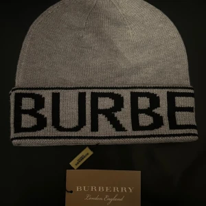 Burberry Mössa - Snygg grå stickad mössa från Burberry med bred svart logotyp runt hela kanten. Klassisk form och mjukt material som håller dig varm. Perfekt för dig som vill ha en stilren accessoar med lyxig känsla och tydlig branding.