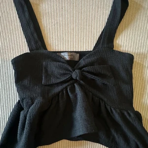 Svart linnetopp med rosett från Primark - Supersöt svart linnetopp från Primark i storlek S. Toppen har breda axelband, en stor rosett framtill och volang nedtill. Materialet är ribbad polyester som ger en cool struktur. Perfekt att styla med jeans eller kjol för en trendig look.