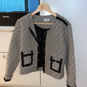 Mönstrad cardigan jacka från By Jolima - Säljer en svartvit cardigan jacka från By Jolima med rutigt diamantmönster, dekorativa knappar och svarta detaljer vid fickor och kanter. Jackan är croppad och har lång ärm, perfekt för dig som vill sticka ut med en cool och trendig look.