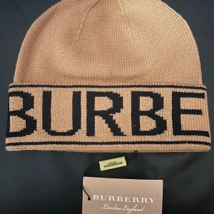 Burberry Mössa - Snygg beige stickad mössa från Burberry med stor svart logotyp runt hela kanten. Klassisk form och mjukt material som håller dig varm. Perfekt för dig som vill ha en lyxig och trendig accessoar med tydlig branding.