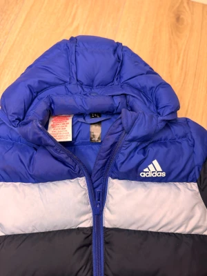 Blå pufferjacka från Adidas 12M - Säljer en blå och svart pufferjacka från Adidas med huva och dragkedja framtill. Jackan har breda färgblock i blått, svart och ljusgrått, samt Adidas-logga på bröstet. Perfekt för kalla dagar och har en cool, sportig vibe.