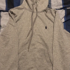 Grå hoodie från Polo Ralph Lauren - Snygg grå hoodie från Polo Ralph Lauren med klassisk logga broderad i mörkblått på bröstet. Tröjan har huva med snörning, stor magficka och långa ärmar. Perfekt för chill dagar och enkel att matcha med jeans eller joggers. Storlek M passar till S och smalla personer, priset kan diskuteras.