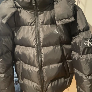 Svart pufferjacka från Calvin Klein Jeans - Säljer en svart pufferjacka från Calvin Klein Jeans med stor CK-logga på ärmen och mindre logga på bröstet. Jackan har dragkedja framtill, hög krage och vadderad design som håller dig varm under kalla dagar. Perfekt för dig som vill ha en stilren och trendig vinterjacka. Pris kan diskuteras vid snabb affär. Skriv vid funderingar🤝