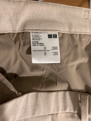 Beige byxor från Uniqlo M - Snygga beige byxor från Uniqlo i storlek M med rak passform och klassiska fickor bak. Materialet är en mix av polyester, viskos och elastan som ger en mjuk och bekväm känsla. Perfekt för dig som gillar stilrena och enkla byxor.