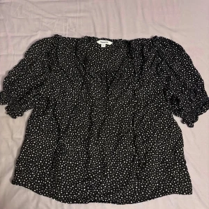 Svart prickig blus från Kappahl - Säljer en svart blus från Kappahl med vita prickar och puffiga korta ärmar. Blusen har en söt volangkant vid halsen och knytband i nacken. Den är i storlek S och inga tecken på användning då den är använd 1 gång.