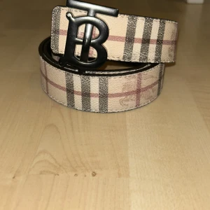 Burberry vändbart skärp med TB-spänne - Säljer ett vändbart Burberry-bälte med ikoniskt TB-spänne i matt svart metall. Ena sidan har klassiskt beige och rutig Burberry-mönster med svarta och röda ränder, andra sidan är svart med diskret rutmönster och ingraverad ryttare. Bältet är tillverkat i skinn och har flera hål för justering.