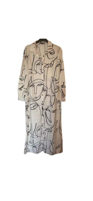 Vit långskjortklänning med ansiktsmönster - Snygg vit lång skjortklänning med svart grafiskt ansiktsmönster. Klänningen har krage, knappar framtill och långa ärmar med manschett. Materialet är mjukt och luftigt.