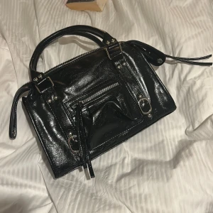 Svart handväska - Snygg svart handväska i glansigt skinn med silverfärgade detaljer och dragkedjor. Väskan har både handtag och en avtagbar axelrem, samt en praktisk ytterficka med dragkedja framtill. HELT ny. Vid frågor kontakta mig!💞