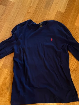 Marinblå långärmad tröja Polo Ralph Lauren - Snygg marinblå långärmad tröja från Polo Ralph Lauren med klassisk röd logobrodyr på bröstet. Tröjan har rund hals och är gjord i mjuk bomull, perfekt för chill dagar. Enkel och stilren design som funkar till jeans eller shorts.
