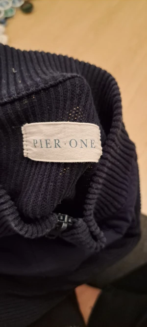 Mörkblå stickad cardigan från Pier One - Snygg mörkblå ribbstickad cardigan från Pier One med rund logopatch framtill. Klassisk form och mjukt material som håller dig varm. Perfekt för kalla dagar och enkel att matcha med din stil.
