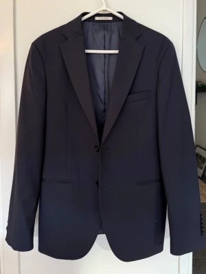 Mörkblå kostym från POUR - Klassisk mörkblå kostym från POUR med stilren kavaj och matchande kostymbyxor. Kavajen har två knappar, smala slag och fickor med lock. Byxorna är raka med pressveck och bälteshällor. Perfekt för dig som vill ha en clean och snygg look.