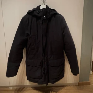 Navyblå J.lindeberg Scandinavian Parka  - Navyblå Scandinavian Parka från J.Lindeberg med dunfyllning och vaxad yta som gör den vattenavvisande och vindtät. Jackan har huva med snörning, stora fickor framtill och snygg patch på ärmen. Perfekt för kalla dagar och stilren look.