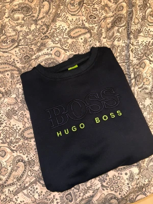 Svart sweatshirt från Hugo Boss - Svart sweatshirt från Hugo Boss med stor broderad logga i mörkblått och neongrönt på bröstet. Tröjan har rund hals och ribbade muddar. Materialet känns mjukt och är troligtvis bomull eller polyester. Perfekt för dig som gillar stilrena och snygga märkesplagg. Står att den är storlek XXL men sitter som L