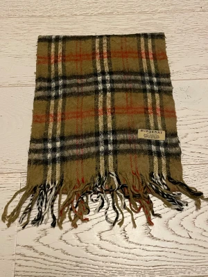 Rutig halsduk från Burberry i ull - Klassisk rutig halsduk från Burberry i 100% lammull. Beige bas med svarta, vita och röda rutor, samt fransar i ändarna. Perfekt accessoar för att lyfta din outfit med ikonisk brittisk stil.