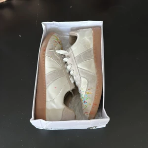 Maison Margiela sneakers med färgstänk - Säljer ett par Maison Margiela sneakers i beige och vitt skinn och mocka med unika färgstänk i grönt, rött, blått och gult. Skorna har klassisk gummisula, snörning och rund tå. Kommer i originalkartong med Margielas ikoniska siffror på locket.  Det är inte original och därför inte lika stora som de egentligen ska vara alltså 43. 