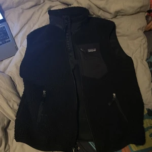Svart fleeceväst från Patagonia - Svart fleeceväst från Patagonia med hög krage och dragkedja framtill. Utsidan är fluffig och mjuk, med två sidofickor med dragkedja och en bröstficka med logga. Perfekt för lager-på-lager och chill stil.