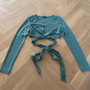 Grön croppad omlottopp från SHEIN - Snygg grön croppad topp från SHEIN med långa ärmar och omlottknytning i midjan. Toppen har v-ringning och en liten öppning framtill som ger en cool detalj. Mjuk och stretchig polyester som sitter skönt mot kroppen. Perfekt för dig som gillar trendiga looks.