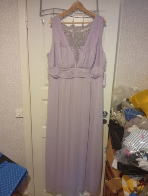 Ljuslila långklänning med pärlor - Långklänning i ljuslila från Bridal's Access, med v-ringad front och vacker pärlbroderad mesh vid halsen. Klänningen är ärmlös, har veckad midja och ett skirt, flytande tyg som ger en elegant siluett. Perfekt för fest och bröllop. Storlek är plus size 22w motsvarar ca 2/3xl