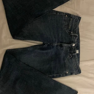 Mörkblå lågmidjade bootcut jeans - Snygga mörkblå jeans med klassisk femficksdesign och bootcut-ben. Jeansen har en stilren look med silverfärgade detaljer vid knappar och nitar. Passar dig som gillar bredare benslut och en tidlös jeansstil.