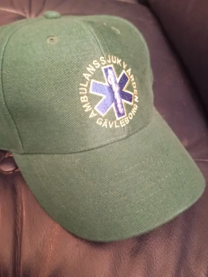Keps Ambulanssjukvården Gävleborg - Snygg grön keps med broderad blå stjärna och gul text 'Ambulanssjukvården Gävleborg' framtill. Klassisk böjd skärm och ventilerande hål upptill. Materialet känns som bomull och kepsen har en normal passform. Perfekt för dig som gillar unik och officiell merch.