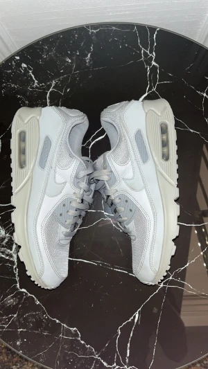 Nike Air Max 90 grå/vita sneakers - Fräscha Nike Air Max 90 sneakers i gråvitt med mesh och skinnpaneler. Klassisk chunky sula med synlig Air-enhet och snygga detaljer. Rund tå, snörning och platt sula gör dem både stilrena och bekväma. Perfekta för dig som gillar att kombinera det ihop med jeans, mjukis eller något överlag. 