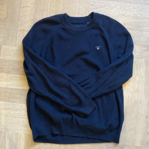 Mörkblå stickad tröja från GANT - Snygg mörkblå stickad tröja från GANT med ribbad struktur och broderad logga på bröstet. Rund hals och långa ärmar ger en klassisk look. Perfekt för dig som gillar stilrena och tidlösa plagg med premiumkänsla.