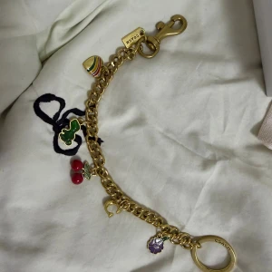 Coach guldigt charmarmband med berlocker - Coach armband i guldfärgad metall med chunky kedja och flera färgglada berlocker: regnbågshjärta, grön björn, röda körsbär, lila blomma och ett litet C. Perfekt accessoar för dig som gillar lekfulla detaljer och exklusiv känsla.