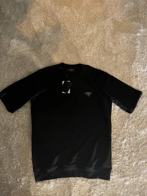 Svart långärmad tröja från Prada - Svart långärmad tröja från Prada med diskret logotyp på bröstet. Tröjan har en klassisk rund hals och är gjord i ett mjukt, glansigt material som ger en lyxig känsla. Perfekt för dig som gillar stilrena och exklusiva plagg.