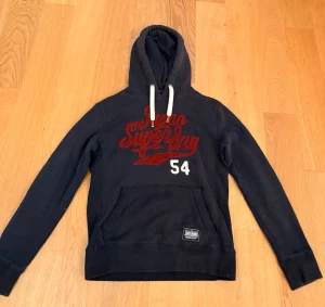 Mörkblå Superdry hoodie med tryck - Snygg trendig mörkblå hoodie från Superdry med rött broderat tryck och vita detaljer. Tröjan har huva med vita snören, stor magficka och långärmade ärmar. 