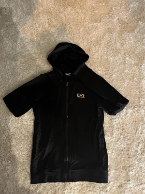 Svart EA7 hoodie  - Svart hoodie från EA7 Emporio Armani. Tröjan har dragkedja framtill och en gul EA7-logga på bröstet. Perfekt för dig som gillar sportig och stilren look. Materialet känns mjukt och har en lätt glansig finish.