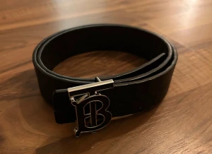 Svart Burberry bälte - Säljer ett svart Burberry bälte i skinn med klassiskt rutigt mönster och präglad riddare. 
