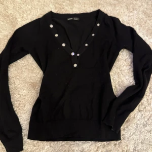 Svart v-ringad tröja med knappar - Svart långärmad tröja SHEIN T  med v-ringning och silvriga knappar längs halsen. Tröjan har en slim passform och är tillverkad i mjuk bomull. 