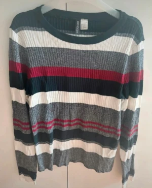 Randig ribbad tröja från H&M - Säljer en snygg långärmad tröja från H&M med breda ränder i vitt, grått, mörkblått och rött. Tröjan är ribbad och har en rund halsringning. Perfekt för dig som gillar färg och mönster i din garderob
