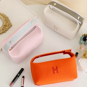 Färgglada handväskor med dragkedja - Tre snygga handväskor i orange, rosa och beige med stilren H-brodering framtill. Väskorna har praktiskt handtag och dragkedja upptill, perfekta för smink eller småprylar. Materialet är mjukt och känns som bomull, med kontrastfärgade detaljer på handtaget.