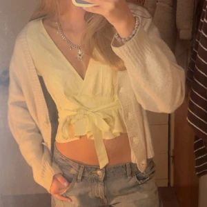 Ljusgul omlottopp med knyt - Supersöt ljusgul omlottopp med knyt i midjan och v-ringad hals. Toppen är croppad och har volangdetaljer nedtill. Perfekt för vår och sommar, och den är ärmlös för en fräsch look. Passar dig som gillar mjuka färger och trendiga detaljer.