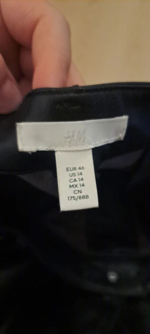 Svarta raka byxor i skinnimitation H&M - Snygga svarta byxor från H&M i storlek 46, tillverkade i skinnimitation med raka ben och klassisk femficksdesign. Byxorna har bälteshällor och knappstängning framtill, perfekt för dig som gillar en stilren look.