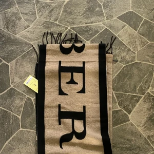 Beige och svart halsduk från Burberry - Snygg och lång halsduk från Burberry med stor logotyp i beige och svart. Halsduken har fransar i ändarna och en bred, rektangulär form. Perfekt för att ge din outfit en lyxig touch. Materialet är mjukt och känns riktigt skönt mot huden.